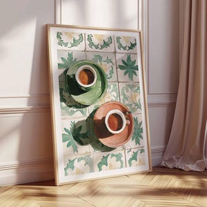 May include: A framed art print featuring two coffee cups on saucers. One cup is on a green saucer, the other on a peach saucer. The background is a tile pattern with green and yellow floral designs. The print is leaning against a white wall and wooden floor.