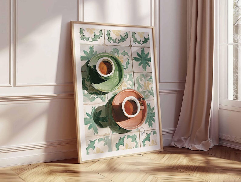 May include: A framed art print featuring two coffee cups on saucers. One cup is on a green saucer, the other on a peach saucer. The background is a tile pattern with green and yellow floral designs. The print is leaning against a white wall and wooden floor.