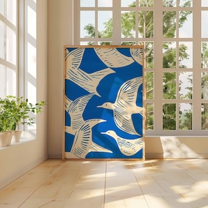 May include: A framed art print featuring a blue background with a pattern of white birds in flight. The artwork is displayed in a room with natural light, near a window and potted plants. The frame is a light brown color.