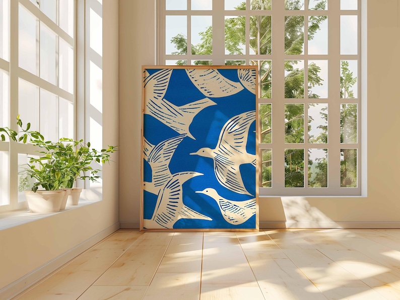 May include: A framed art print featuring a blue background with a pattern of white birds in flight. The artwork is displayed in a room with natural light, near a window and potted plants. The frame is a light brown color.