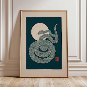 May include: Framed art print depicting a teal and gold snake coiled in front of a full moon on a dark teal background. The artwork has a minimalist style with a red stamp in the lower right corner.
