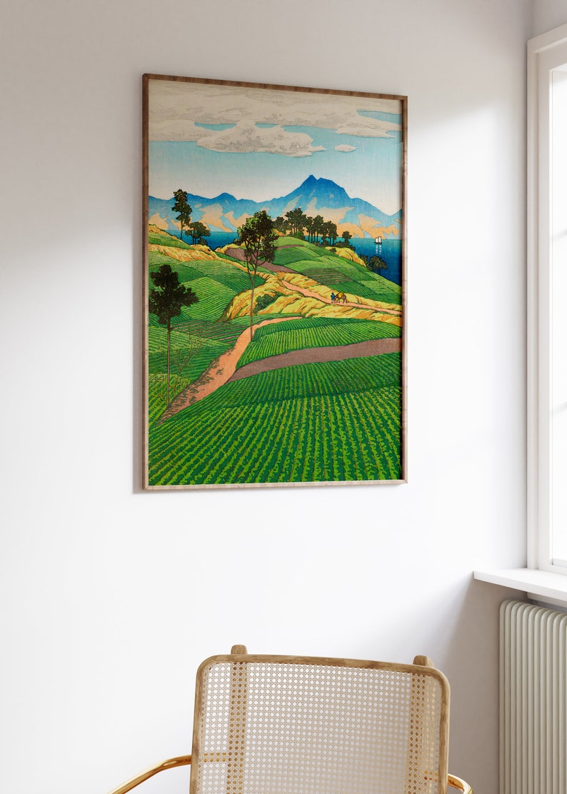 May include: A framed woodblock print of a Japanese landscape. The print depicts a winding road leading up a hillside with green fields and trees. In the distance, there are mountains and a body of water. The sky is blue with white clouds.