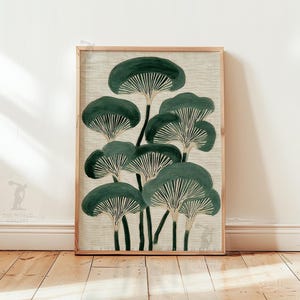 May include: A framed art print featuring a stylized illustration of dark green mushrooms with white gills against a beige background. The artwork is displayed in a light brown wooden frame, leaning against a white wall and wooden floor.