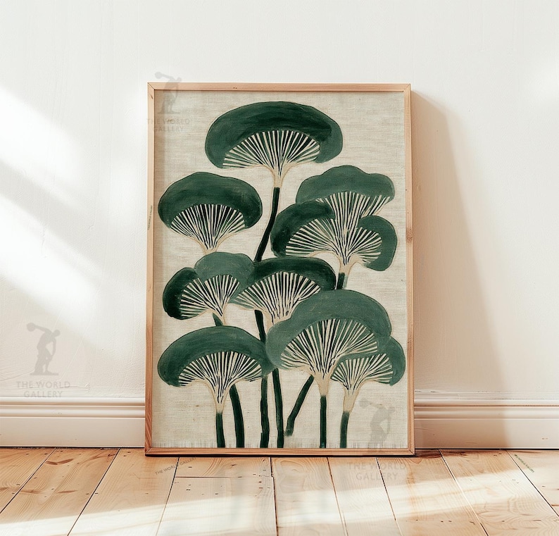 May include: A framed art print featuring a stylized illustration of dark green mushrooms with white gills against a beige background. The artwork is displayed in a light brown wooden frame, leaning against a white wall and wooden floor.