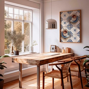 May include: A wooden table with a light wood finish and metal legs. Two chairs with woven seats and backs are positioned at the table. A window with white trim and a view of trees is in the background. A silver pendant light hangs above the table. A framed print with a blue and white tile pattern and a coffee cup design is on the wall.