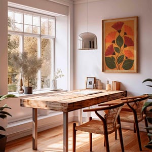 May include: A bright, airy room with a wooden desk and two chairs. A framed floral print hangs on the wall. A large window offers a view of trees. A silver pendant light hangs above the desk. The room is decorated with plants and other decor.