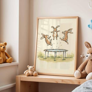 May include: A framed illustration depicts three brown rabbits jumping on a trampoline. The artwork is set against a light background, with a wooden frame. The scene is reminiscent of a children's book illustration, with a playful and whimsical theme.