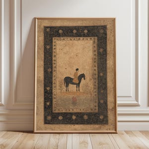 May include: Framed vintage artwork depicting a person on horseback. The artwork has a beige background with a black floral border. The person is wearing a tan outfit and riding a black horse. The frame is a light brown colour.