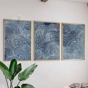 May include: Three framed wall art pieces with a navy blue and white wave pattern. The artwork is encased in gold frames and mounted on a white wall. A green plant is visible in the foreground.