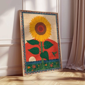 Op de afbeelding: Een ingelijste kunstdruk met een grote gele zonnebloem met een bruin centrum, groene bladeren en een rode en oranje zonsondergangachtergrond. Een kolibrie is ook zichtbaar. Het kunstwerk leunt tegen een witte muur en een houten vloer.