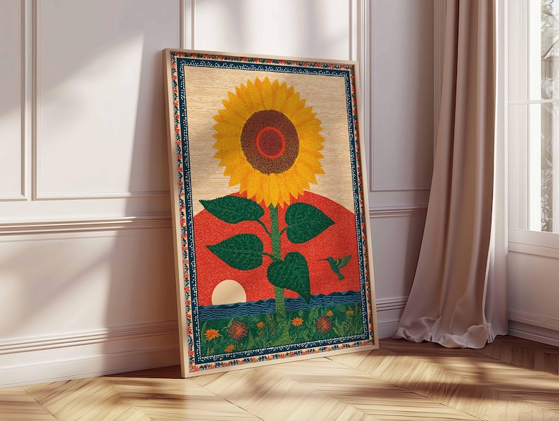 Op de afbeelding: Een ingelijste kunstdruk met een grote gele zonnebloem met een bruin centrum, groene bladeren en een rode en oranje zonsondergangachtergrond. Een kolibrie is ook zichtbaar. Het kunstwerk leunt tegen een witte muur en een houten vloer.