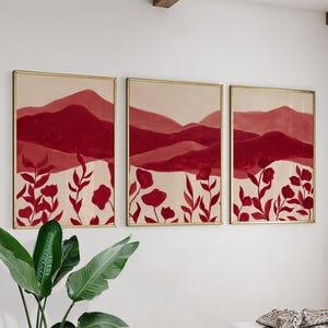 May include: Set of three framed art prints. Each print features a mountain landscape in shades of red and burgundy, with floral accents in the foreground. The frames have a gold finish.
