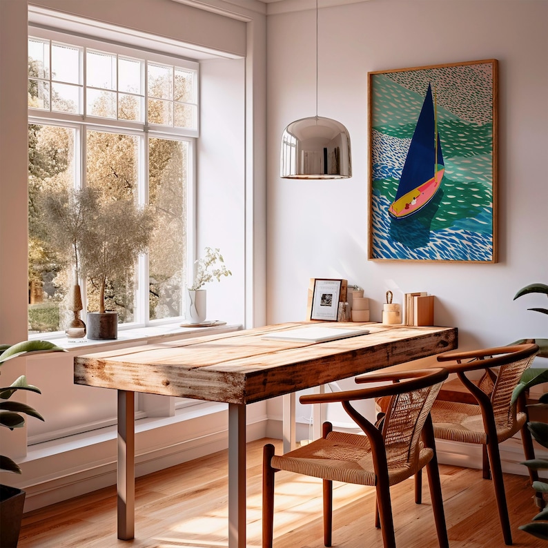 May include: A light and spacious room featuring a large window with a view of trees. A wooden desk with metal legs and two woven chairs are positioned in front of the window. A framed sailboat art print adorns the wall, and a chrome pendant light hangs from the ceiling.