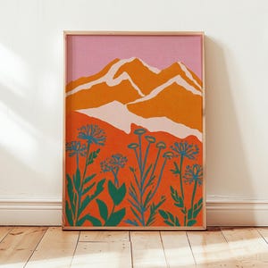 以下が含まれることがあります： オレンジ色の山頂とピンク色の空が描かれた、額入りのアートプリント。前景には、オレンジ色の背景に様式化された青い花と緑の葉が描かれています。アートワークは木製のフレームに入っています。