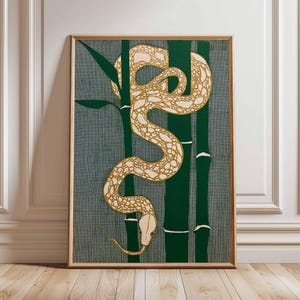 May include: A framed art print depicting a cream and gold snake entwined around green bamboo. The background is a textured grey-blue. The artwork is presented in a wooden frame, suitable for wall display.