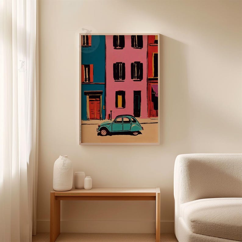 May include: A framed art print depicts a colorful street scene with buildings in teal and pink. A teal vintage car is parked in front of the buildings. The artwork is displayed on a beige wall above a wooden table with white vases.