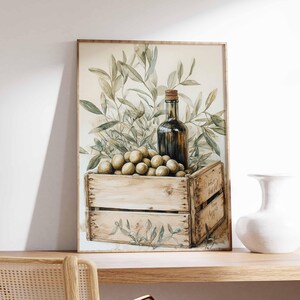 May include: Watercolor painting of olives, olive branches, and a bottle of olive oil in a wooden crate. The artwork is framed in a light brown frame and is set on a wooden shelf. A white vase is to the right of the painting.