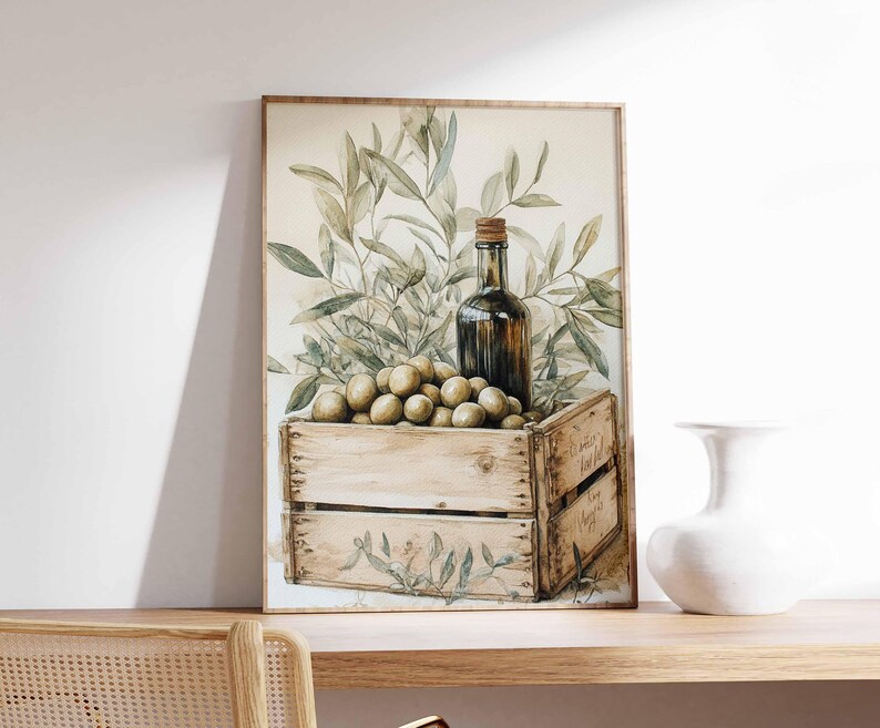 May include: Watercolor painting of olives, olive branches, and a bottle of olive oil in a wooden crate. The artwork is framed in a light brown frame and is set on a wooden shelf. A white vase is to the right of the painting.