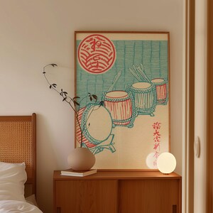 May include: A framed print featuring Japanese drums in red and teal against a light blue background. The artwork includes a red circular emblem and Japanese text. The print is displayed above a wooden cabinet with a vase and a lamp.
