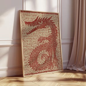 May include: A framed print featuring a red dragon illustration. The dragon is depicted with sharp teeth and claws, set against a textured beige background. The artwork is displayed in a light-colored wooden frame, leaning against a white wall and wooden floor.