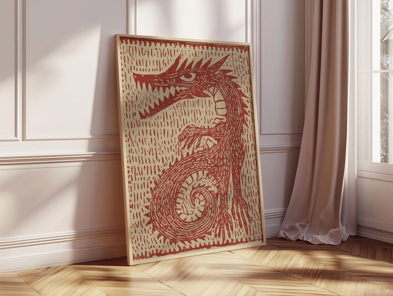 May include: A framed print featuring a red dragon illustration. The dragon is depicted with sharp teeth and claws, set against a textured beige background. The artwork is displayed in a light-colored wooden frame, leaning against a white wall and wooden floor.