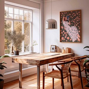 K&ouml;nnte beinhalten: Ein heller, luftiger Raum mit einem Holztisch und zwei St&uuml;hlen. Ein gro&szlig;es Fenster bietet einen Blick auf B&auml;ume. Ein gerahmtes Kunstwerk zeigt eine Person, die in einem Blumenfeld liest. Eine silberne Pendelleuchte h&auml;ngt &uuml;ber dem Schreibtisch.