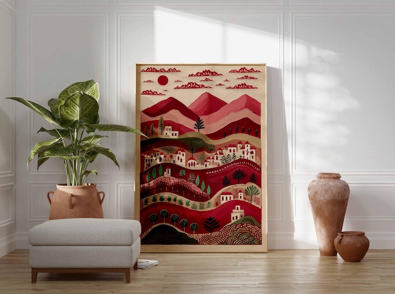May include: A framed art print showing a stylized landscape in shades of red, pink, and green. The artwork features a village with white buildings, trees, and a red sun. The print is displayed with a large potted plant, a footstool, and decorative vases.