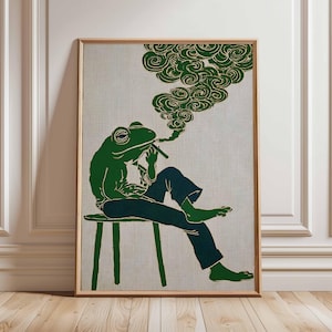 May include: A framed art print depicting a green frog smoking a cigar, seated on a stool. The frog is wearing a hat and dark blue trousers. Green smoke swirls above the frog's head. The background has a neutral linen texture.