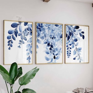 May include: Set of three framed art prints featuring watercolor paintings of blue leaves and branches against a white background. The frames are gold-colored, adding a touch of elegance to the botanical artwork.