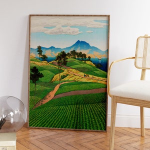 May include: A framed print of a Japanese landscape painting depicting a winding road leading up a hill with green fields and trees. In the distance, there are mountains and a body of water with a sailboat.