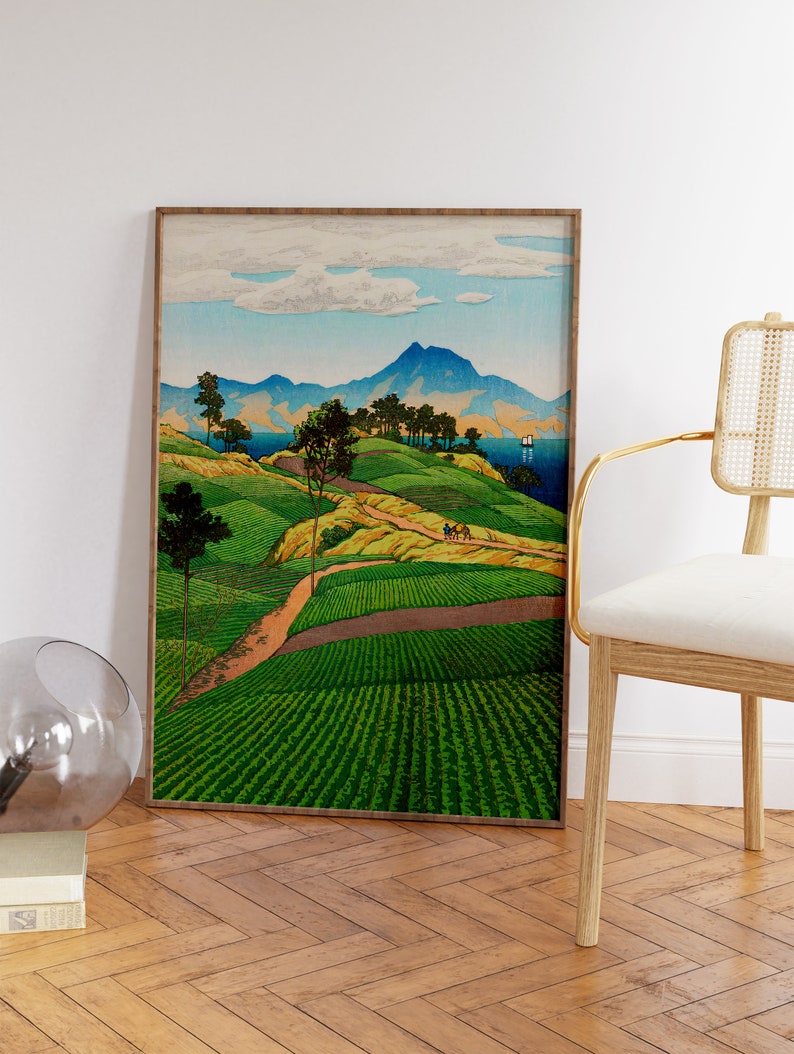 May include: A framed print of a Japanese landscape painting depicting a winding road leading up a hill with green fields and trees. In the distance, there are mountains and a body of water with a sailboat.