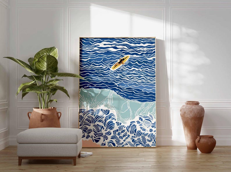 May include: A framed art print depicting a surfer on a surfboard in the ocean. The artwork features shades of blue and white to represent the water and waves. The print is displayed in a room with a plant, ottoman, and decorative vases.