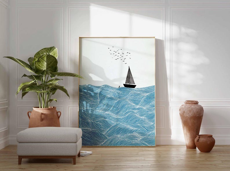 May include: A framed art print featuring a sailboat on a wavy blue sea, with birds in the sky. The artwork is displayed in a room with a large potted plant, a gray ottoman, and decorative vases. The frame has a gold finish.