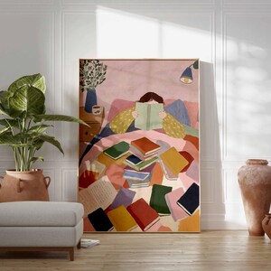 May include: A framed art print depicts a person reading in bed, surrounded by colorful books. The artwork features a warm color palette with pinks, yellows, and blues. A large potted plant, a gray ottoman, and terracotta vases are in the foreground.