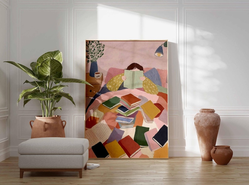 May include: A framed art print depicts a person reading in bed, surrounded by colorful books. The artwork features a warm color palette with pinks, yellows, and blues. A large potted plant, a gray ottoman, and terracotta vases are in the foreground.