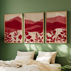 May include: Three framed art prints with a nature theme. Each print features a red and cream color palette with abstract mountain and floral designs. The frames are gold-colored, and the artwork is displayed on a green wall above a bed.