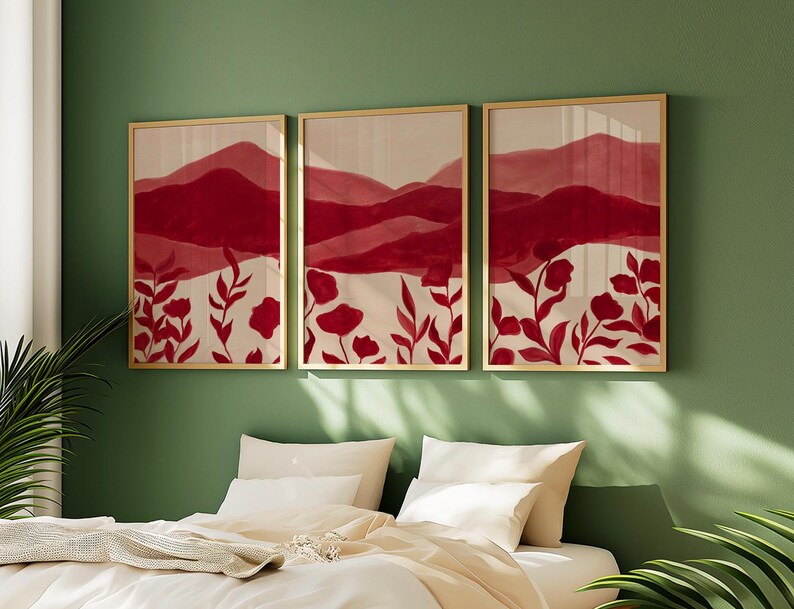 May include: Three framed art prints with a nature theme. Each print features a red and cream color palette with abstract mountain and floral designs. The frames are gold-colored, and the artwork is displayed on a green wall above a bed.