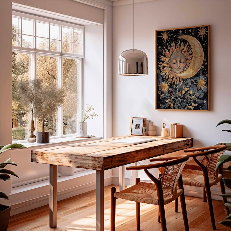 May include: A wooden table with a light wood finish and a metal frame. The table has a large, round, silver pendant light hanging above it. There is a framed print of a sun and moon with a face on the wall behind the table. Two chairs with woven seats are placed at the table.