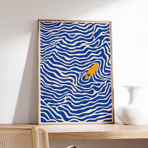 May include: Abstract art print featuring a blue and cream wavy pattern with an orange surfboard floating in the center. The artwork is framed in a natural wood tone and displayed on a wooden shelf. A white vase sits to the right.