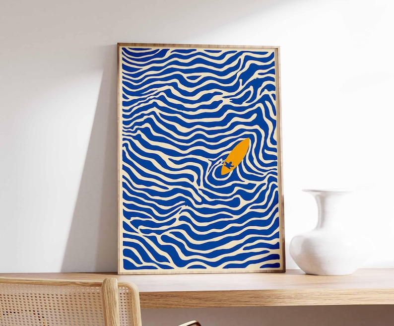 May include: Abstract art print featuring a blue and cream wavy pattern with an orange surfboard floating in the center. The artwork is framed in a natural wood tone and displayed on a wooden shelf. A white vase sits to the right.
