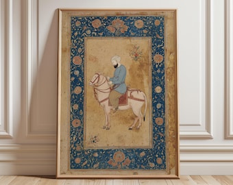 May include: A framed vintage artwork showing a figure on horseback. The artwork features a central image of a person in blue attire riding a pale horse. A decorative blue border with floral patterns surrounds the central image. The frame is a warm, light brown.
