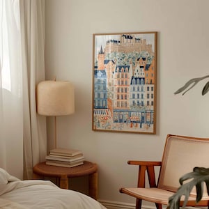 May include: A framed art print showing a European town with colorful buildings, a castle, and a street scene with people. The artwork is in a wooden frame and hangs on a neutral wall. The room also features a lamp, books, and a chair.