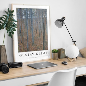 May include: A framed print of a painting by Gustav Klimt titled "Beech Grove I, 1902". The painting depicts a forest scene with trees and leaves in shades of blue, green, and brown. The print is displayed on a desk with a laptop, a lamp, and other office supplies.