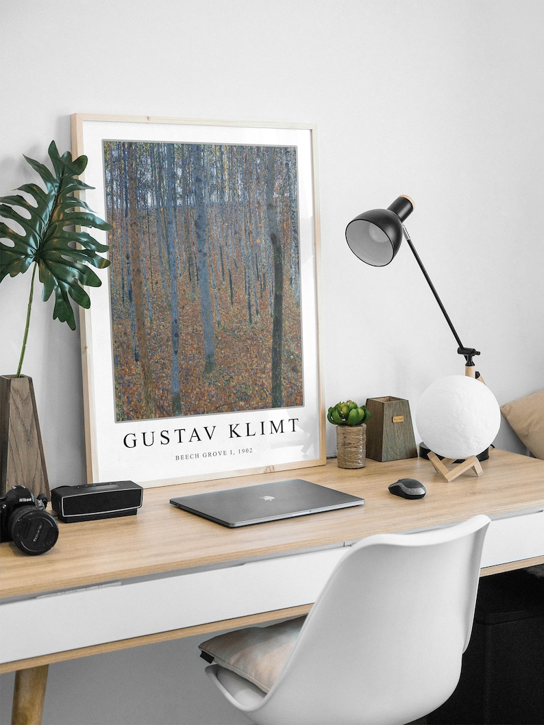 May include: A framed print of a painting by Gustav Klimt titled "Beech Grove I, 1902". The painting depicts a forest scene with trees and leaves in shades of blue, green, and brown. The print is displayed on a desk with a laptop, a lamp, and other office supplies.