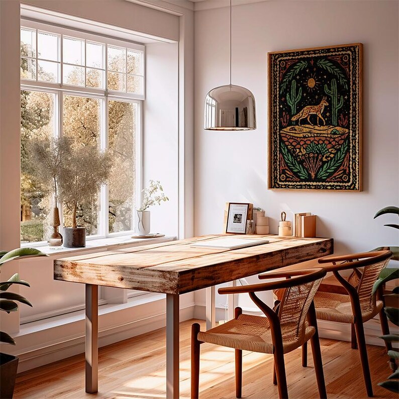 May include: A rustic wooden desk with metal legs and two woven chairs sits in front of a large window. A framed desert-themed tapestry with a fox, cacti, and sun hangs on the wall. A silver pendant light hangs above the desk.