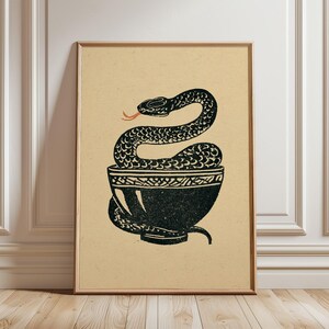 Op de afbeelding: Een ingelijste kunstdruk op een lichtbruine achtergrond met een zwarte inktillustratie van een slang die in en rond een donkere kom is gekronkeld. De slang heeft een zichtbare rode tong en de kom heeft een decoratief patroon langs de rand.