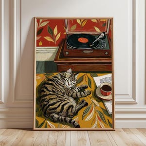 Könnte beinhalten: Gerahmter Kunstdruck, der eine getigerte Katze zeigt, die auf einer gelb gemusterten Oberfläche schläft. Im Hintergrund befindet sich ein Vintage-Plattenspieler mit einer Schallplatte auf dem Plattenteller. Eine Tasse Kaffee steht daneben und schafft eine entspannte, gemütliche Szene.