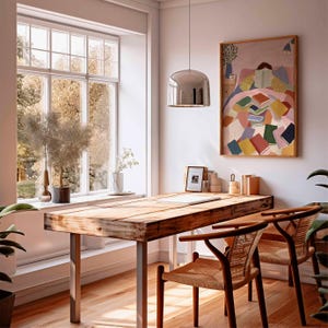 May include: A bright, airy room with a wooden desk and two chairs. A large window offers a view of trees. A colorful painting of a person reading sits above the desk. A silver pendant light hangs from the ceiling.