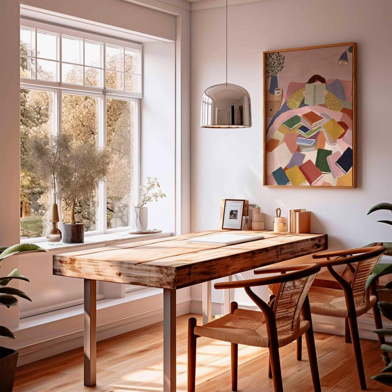 May include: A bright, airy room with a wooden desk and two chairs. A large window offers a view of trees. A colorful painting of a person reading sits above the desk. A silver pendant light hangs from the ceiling.