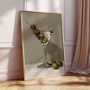 May include: A framed art print depicts a martini glass filled with clear liquid and green olives. The artwork is set against a muted green background, with the glass resting on a white surface. The print is displayed in a room with a wooden floor and a beige curtain.